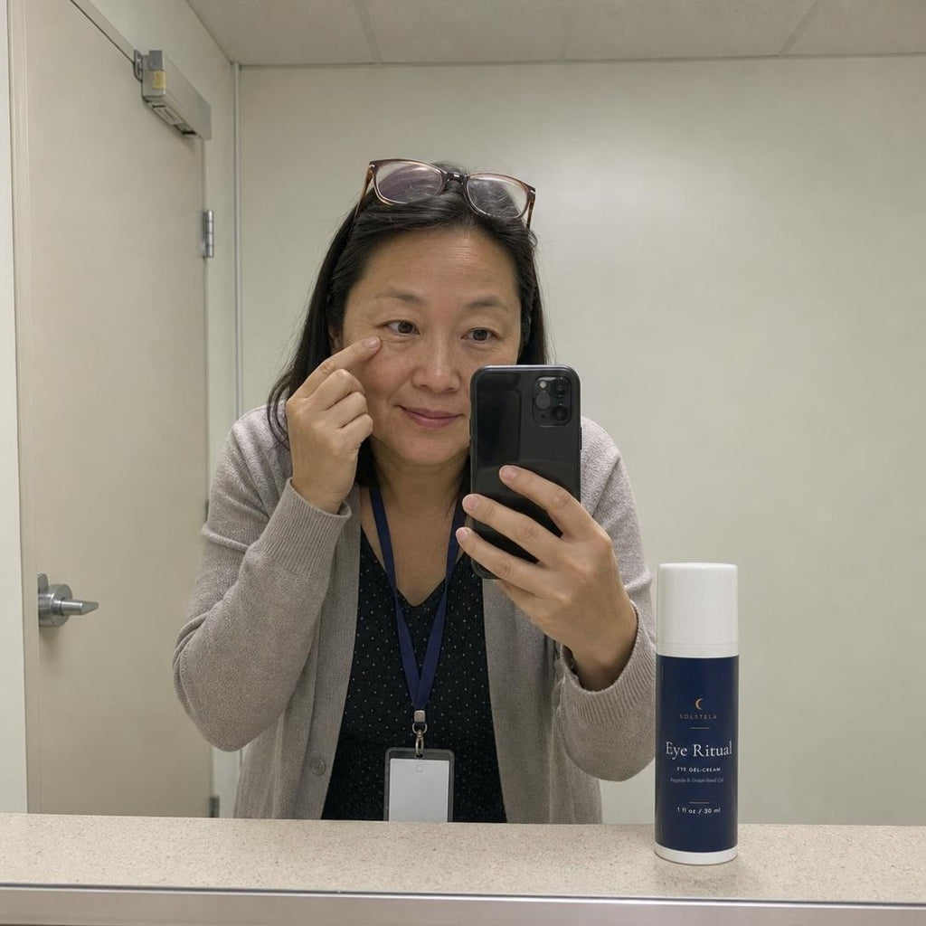 A woman in a cardigan and lanyard takes a mirror selfie in a staff room, fingertip under her eye, with Eye Ritual on the ledge