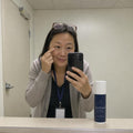 A woman in a cardigan and lanyard takes a mirror selfie in a staff room, fingertip under her eye, with Eye Ritual on the ledge